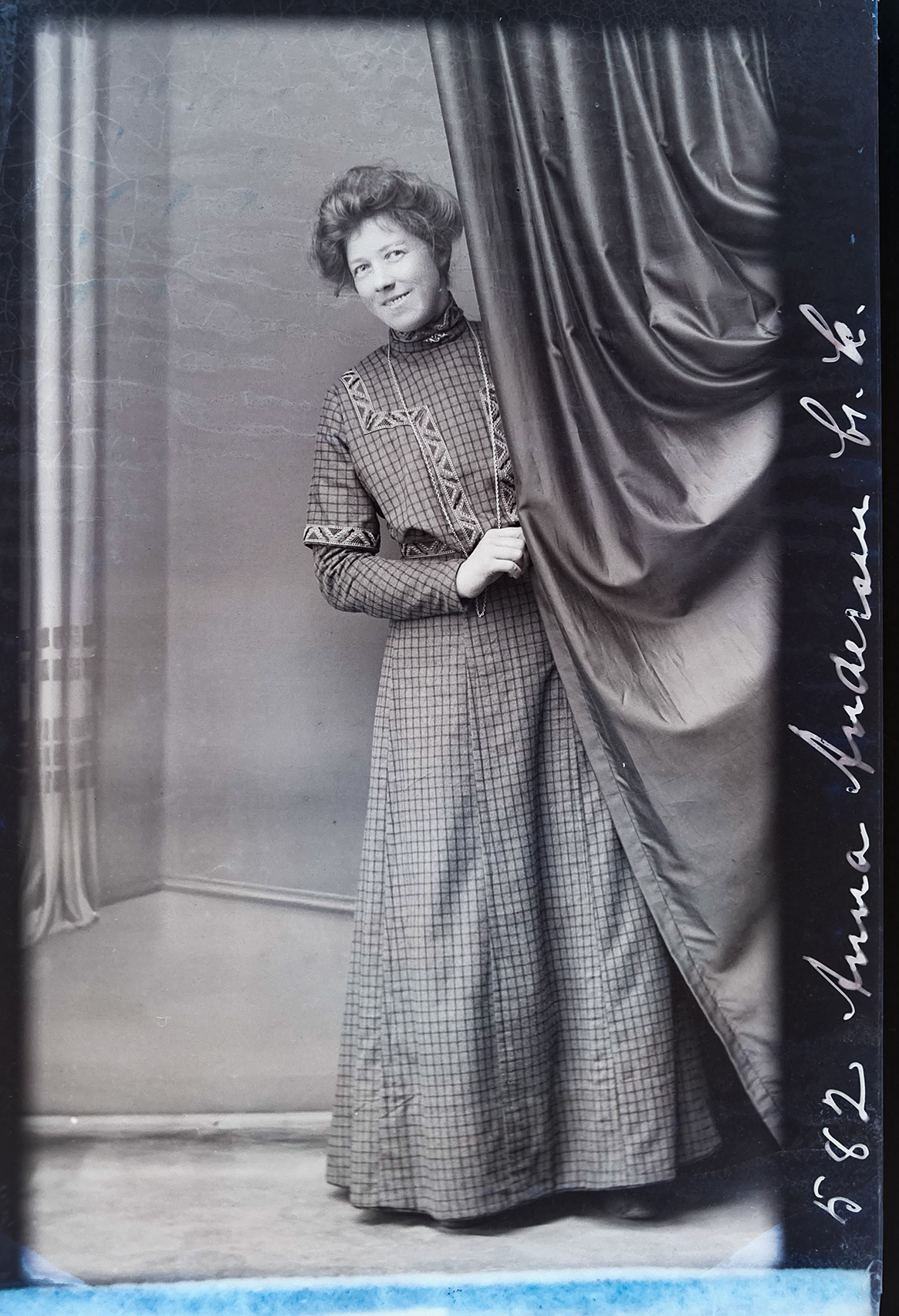 Gammelt svarthvitt fotografi av kvinne i fotsid kjole som titter frem bak gardin.