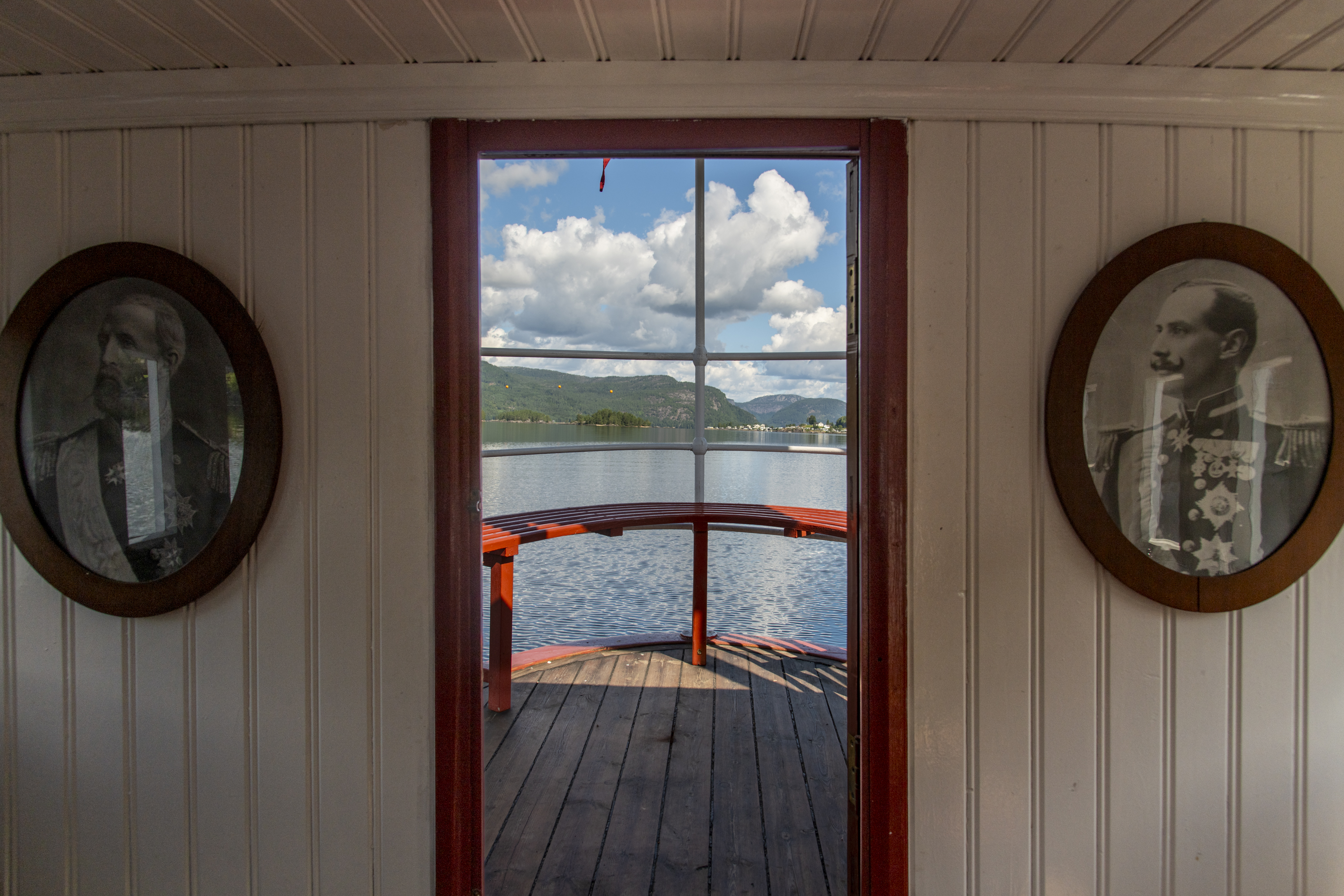 Interiørbilde fra gammelt dampskip. Bildet viser to vegger med kongeportretter og utsikt mot sjøen.
