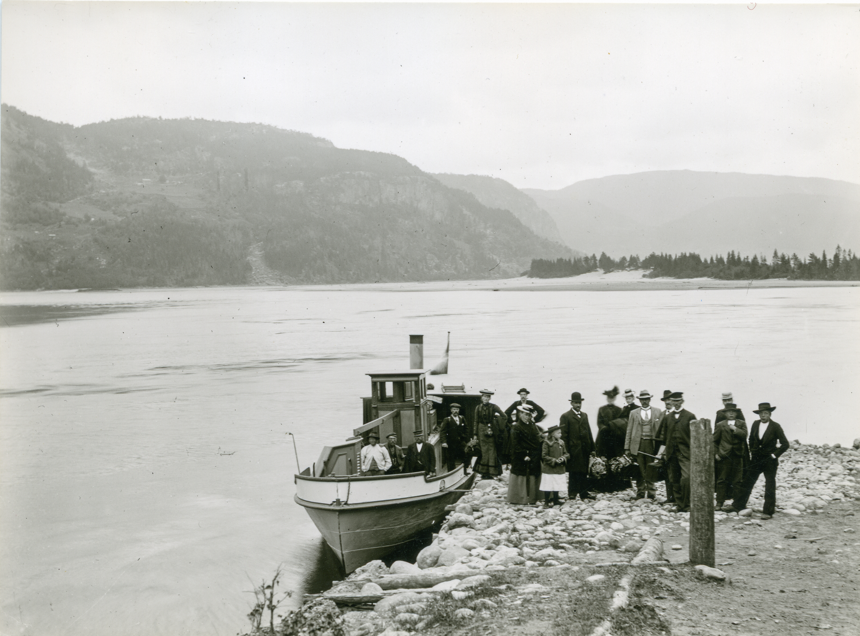 gammelt sort/hvitt -bilde som viser lite dampskip med folk på kai med fjell rundt.