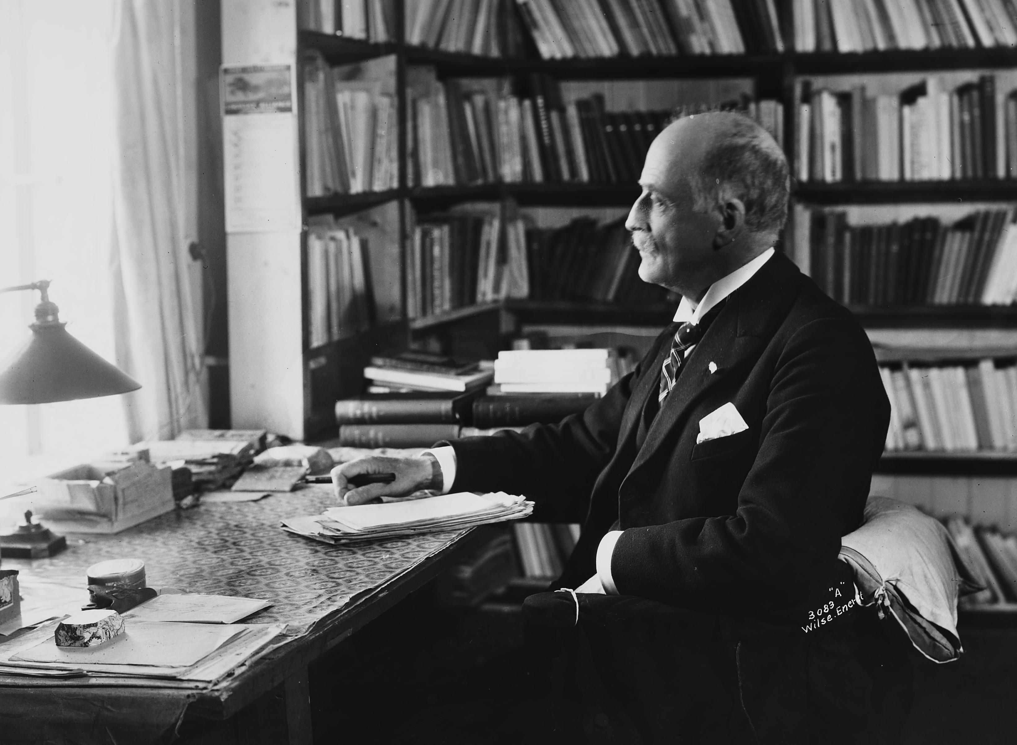 Sorthvittbilde av mann som ser ut av vindu omgitt av bøker. 