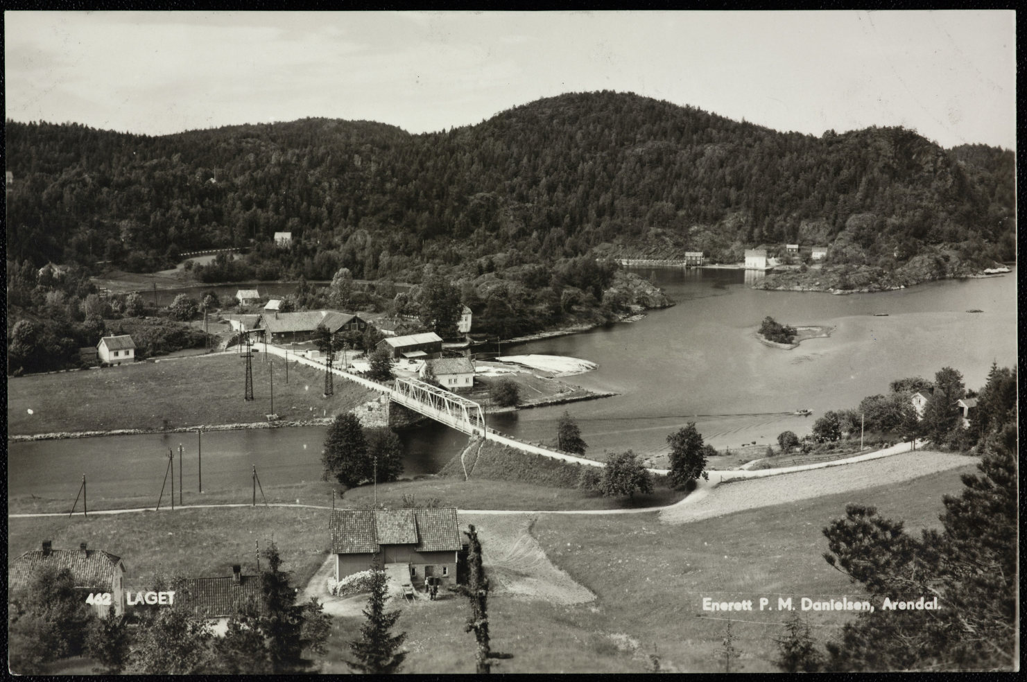 Gammelt svart/hvitt foto som viser landskap med hus, elv og bro. Teksten på bildet er "Laget"